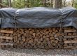 Pile de bois de chauffage fendue, bien rangée sous une bâche grise attachée avec des cordes, dans un environnement boisé.